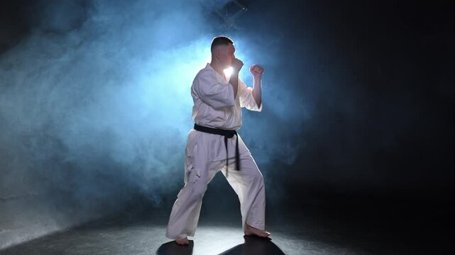 Strong man fighter in white kimono for karate martial arts with black belt preparing for fight training in black background. Man training moves punches kicks practicing shadow fight ready for sparring