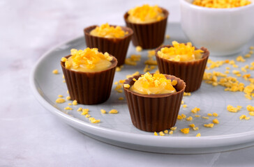 Chocolate cups with open mango filling and orange crunch sprinkles on a ceramic plate on a light background. Holiday dessert. Handmade candies