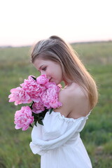 Fototapeta premium A young woman in a white dress stands in a field at sunset, holding a bouquet of pink peonies. Her serene gaze and gentle expression convey a sense of peace and harmony with nature
