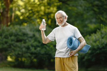 Senior man with exercise mat standing at park and holding bottle of water