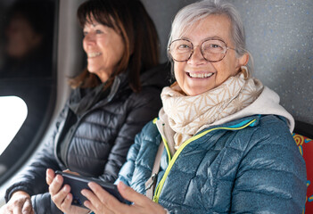 Joyful friendship of two elderly women taking a sightseeing tour on a bus while looking at a cell...