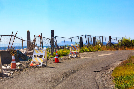 Shoreline erosion, damage to a coastal road on Lummi Island, rubble and broken fences on the roadside, with warning cones and signs.