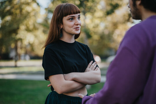 A young couple engages in a friendly conversation outdoors on a sunny day, surrounded by nature. The woman smiles while standing with her arms crossed, conveying a relaxed atmosphere. - Powered by Adobe