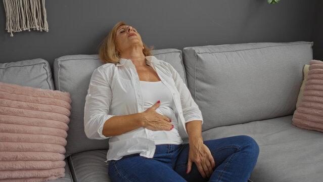 Elderly woman with stomach pain sitting on a sofa in a living room, looking uncomfortable in an indoor home setting.