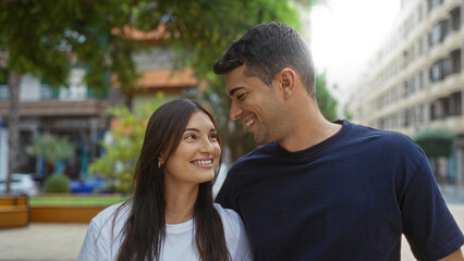 Obraz premium Young couple smiling together in an urban park, with lush trees and city buildings in the background, showcasing love and relationship outdoors in a beautiful setting.