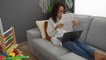 Young woman working from home in living room with laptop and documents on couch surrounded by toys...