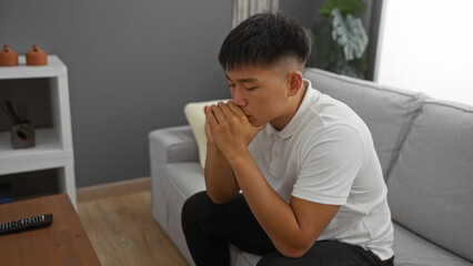 Fototapeta premium Young chinese man sitting indoors in a modern apartment, pondering deeply in the living room, creating a thoughtful and introspective atmosphere