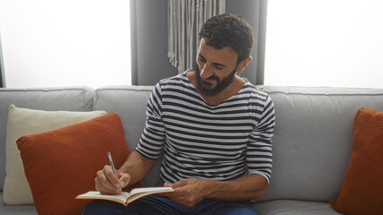 Obraz premium Young man with a beard in a striped shirt writing in a notebook while sitting on a couch at home with orange and white pillows