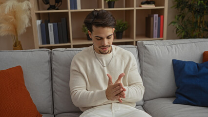 Fototapeta premium Young hispanic man with hands together sits on a cozy sofa in a stylish living room interior, surrounded by modern decor and neutral tones, conveying a relaxed home atmosphere.