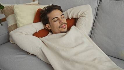 Young hispanic man relaxing on a comfortable sofa indoors, radiating a calm and serene presence in...