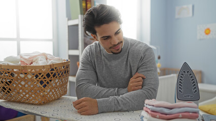 Young man indoors in a laundry room considers his work as he irons clothes and organizes a basket...