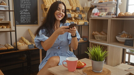 Hispanic woman taking photo of her breakfast with a mobile phone in a cozy bakery cafe, smiling and enjoying the indoor atmosphere with various pastries on display.