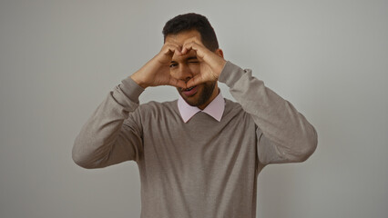 Handsome young hispanic man makes a heart gesture over his face on an isolated white background showcasing emotion and connection