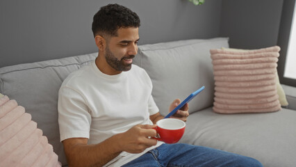 Young man sitting on a couch in a living room holding a red cup and looking at a tablet, dressed in...