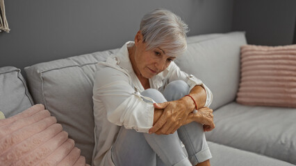 Grey-haired woman sitting thoughtfully on a couch in a cozy living room, conveying a serene and...