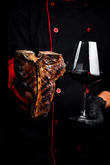 The chef is holding a T-bone steak and a glass of wine. Close-up. On a black background.
