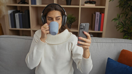 Woman relaxing on sofa with headphones while video chatting on smartphone in cozy apartment,...