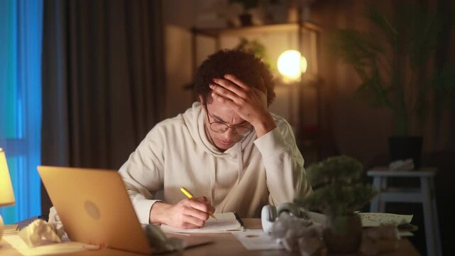 Disappointed young african american man at desk and crossing out ideas in notebook struggling with creative block or frustration while working at home in the evening facing doubt and self-criticism