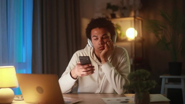 Bored young African American man in headphones staring at smartphone screen at home in the evening after study or work feeling tired distracted or uninterested while browsing or listening to music