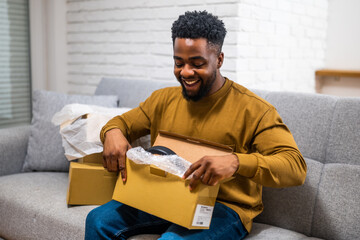 Young African man sitting on cozy sofa in living room and unboxing package with joy after receiving online shopping delivery. Representation of customer satisfaction with home shopping.