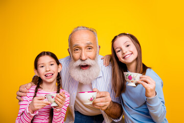 Obraz premium Elderly man enjoying a playful tea party with his two young granddaughters against a vibrant yellow background, showcasing happiness and family bonding