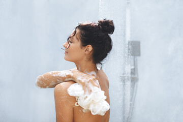 A serene woman with a bun enjoys a refreshing shower, using a foamy loofah to cleanse her skin. The tranquil scene and her calm demeanor highlight the importance of self-care, making it perfect for
