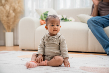 Fototapeta premium Cute baby girl sitting on a plush rug in a cozy living room with natural light, displays curiosity and joy while playing