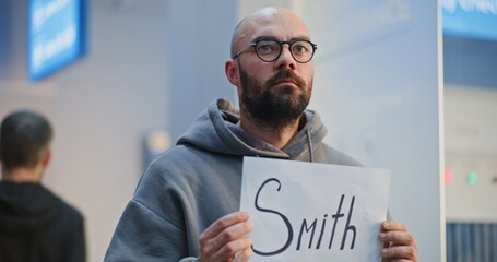 Professional Pickup Service in International Airport Terminal: Airport Greeter Holding Greeting Name Sign, Waiting for Passengers From Plane Flight. Diverse People, Tourists Walking. Medium Close Up. © Framestock