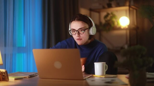 Shocked young woman in headphones watching something on laptop while eating snack at home reacting emotionally to unexpected content during cozy evening in modern relaxed workplace