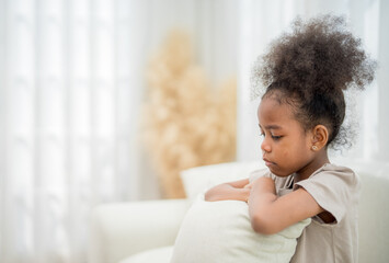 Young girl sitting on a couch with a pillow, deep in thought, in a softly lit room with curtains