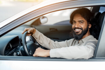 Indian man sitting in new car, smiling confidently at camera, happy handsome eastern male resting arm on window while holding the steering wheel, closeup