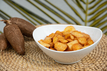 Organic manioca or cassava chips in bowl and tapioca tuber on wicker tray. Indonesia popular hot spicy snacks. Close up.