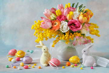 Easter beautiful postcard, bouquet of spring yellow, pink, white flowers in a vase, decorative rabbits, eggs on a blue background