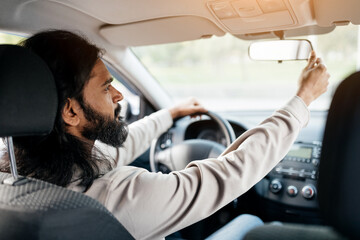 Testing Visibility. Indian Man Adjusting Rear View Mirror, Enjoying New Auto, Sitting On Driver's Seat In Luxury Vehicle. Guy Driving Alone, Over Shoulder View From The Back Passenger Seat