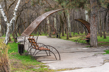 View of four empty park benches with canopies on both sides of the park path in early spring. Birch and pine trees are also in the foreground.