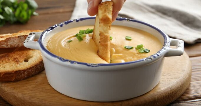 Woman dipping crouton into tasty cheese sauce at wooden table, closeup