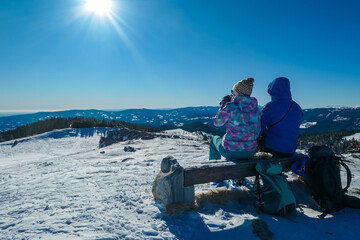 Couple sits on a wooden bench enjoying sunny winter day atop snow-covered W&ouml;lkerkogel in Styria, Austria. Panoramic view of majestic snow-capped mountain peaks in Lavanttal Alps. Snowshoe hiking