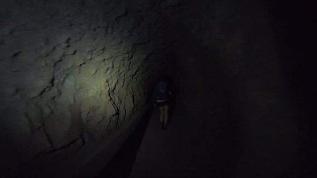 A lone hiker navigates the darkness of a levada tunnel, their silhouette illuminated by faint light from the distant exit ahead.