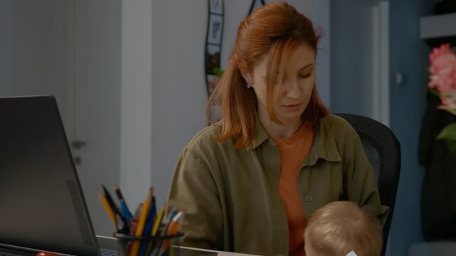 Mother tries to focus on her work at a laptop in a home office while her young child seeks her attention. The challenges of balancing a career and parenting are evident.