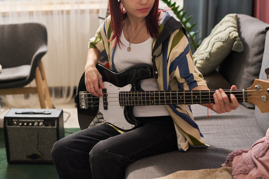 Teenage girl sitting on sofa while playing electric bass guitar in comfortable, serene living room environment. Amplifier and plant visible, creating casual atmosphere