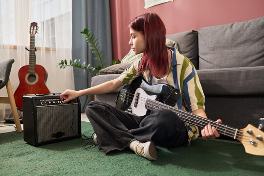 Person seated on carpet, adjusting amplifier controls while holding electric guitar, with acoustic guitar and houseplant in background, showcasing a cozy living space - Powered by Adobe