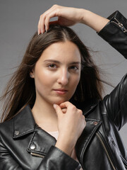 Young woman in a model test, close-up shot. Natural expression, soft smile, and lively emotions. Long hair, leather jacket, neutral background. Visible skin texture, soft studio lighting.