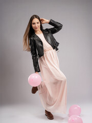 Young woman in a model test, full-length shot. Movement, flowing pink dress, leather jacket—stylish contrast. Neutral background, pink balloons add playfulness and lightness.