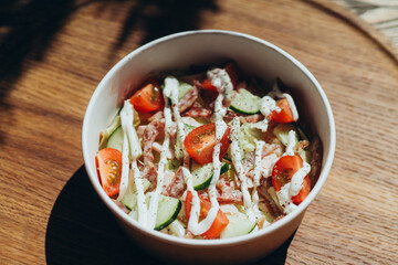 Fresh salad bowl with cherry tomatoes, cucumber, ham and creamy dressing served on wooden tray in bright daylight