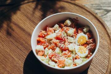 Caesar-style salad with cherry tomatoes, egg halves, croutons and grated cheese in eco-friendly bowl on wooden surface