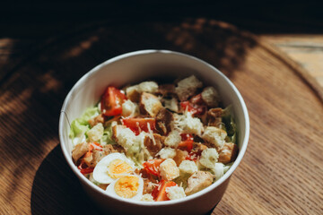 Caesar-style salad with cherry tomatoes, egg halves, croutons and grated cheese in eco-friendly bowl on wooden surface
