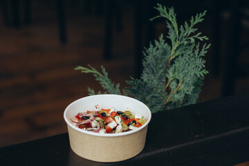 Classic Greek salad with feta cheese, olives and red onion in paper bowl, rustic background with pine