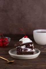 Piece of Black forest cake with cherry, cream with shea butter, cognac and chocolate biscuit and cup of coffee on a brown table. Vegan dessert. Vertical orientation.