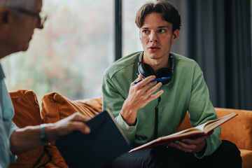 A young man with headphones around his neck converses with an older mentor while holding a book,...