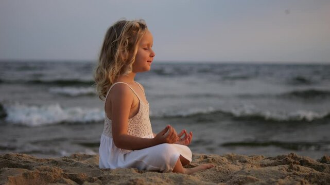 Peaceful blond child practicing yoga on sandy seashore, experiencing serenity during golden sunset with gentle ocean waves surrounding meditative pose. Concept for children meditation